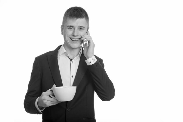 Studio shot of young happy businessman smiling and holding coffee cup while talking on mobile phone - Zdjęcie, obraz