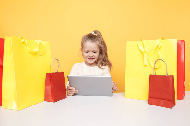 Petite fille souriante en ligne à l'aide d'un ordinateur portable et assis au bureau avec des sacs à provisions. - Photo, image