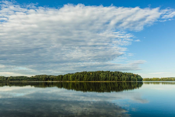 Сентябрьский вечерний пейзаж с голубым небом, белыми облаками, зеленым лесом и его отражением в спокойной воде
. - Фото, изображение