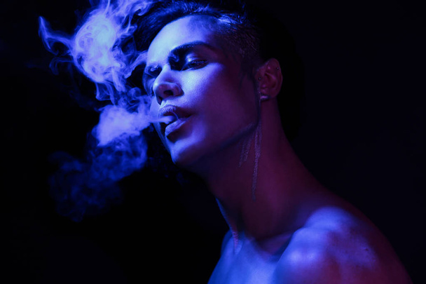 Close up studio portrait of beautiful young man with face art, violet and blue light. Smoking concept  - Foto, afbeelding