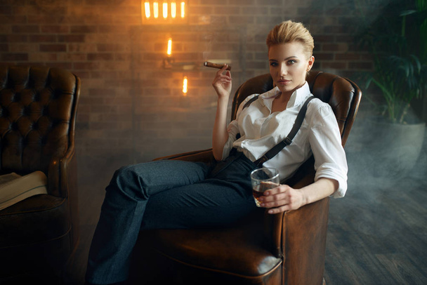 Woman in shirt and trousers sitting in leather chair with whiskey and cigar, retro fashion, gangster style. Vintage business lady in office with brick walls - Fotografie, Obrázek