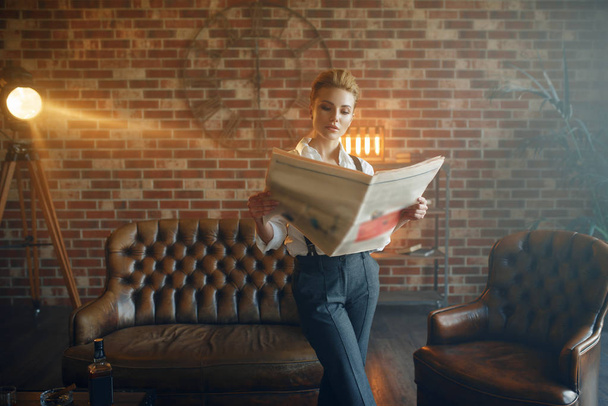 Женщина в рубашке и брюках с подтяжками держит газету, ретро моду, гангстерский стиль. Винтажная деловая леди в офисе с кирпичными стенами
 - Фото, изображение