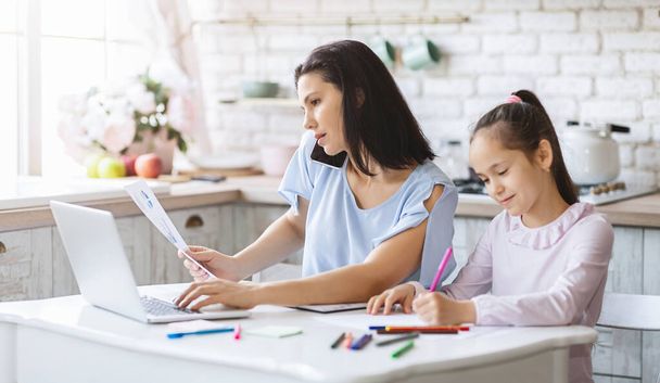 Mother working from home while daughter doing homework in kitchen - Foto, Imagen
