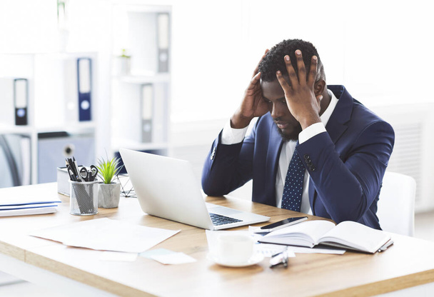 ストレスを受けたアフリカのビジネスマンは、額に手をつないでオフィスに座っている - 写真・画像