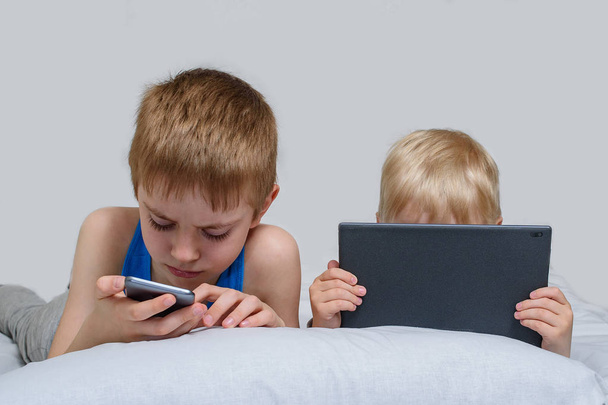 Dos chicos con artilugios yacen en la cama. Los niños usan teléfono inteligente y tableta
 - Foto, imagen