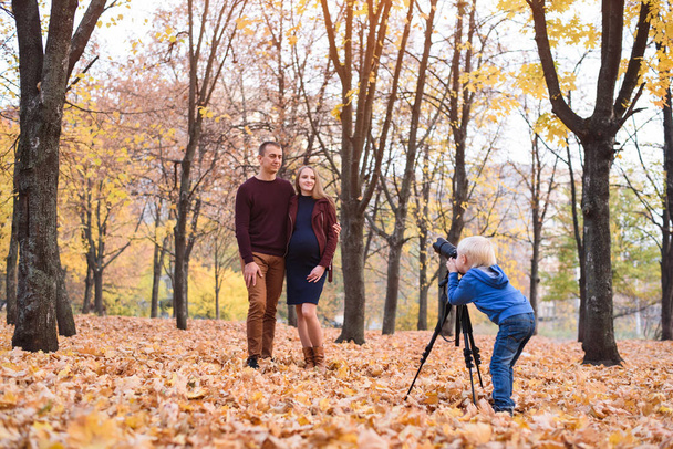 Little blond boy with a big SLR camera on a tripod. Photographs a married couple, pregnancy. Family photo session - Photo, Image