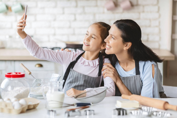 Happy little girl taking selfie with her mother in kitchen. - 写真・画像