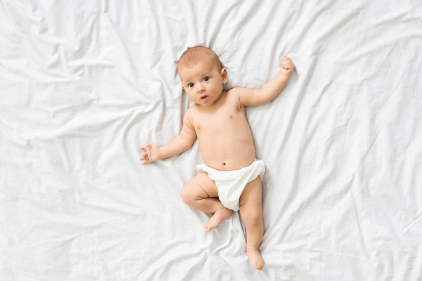 Portrait of cute open-eyed baby in diaper lying on bed - Photo, Image
