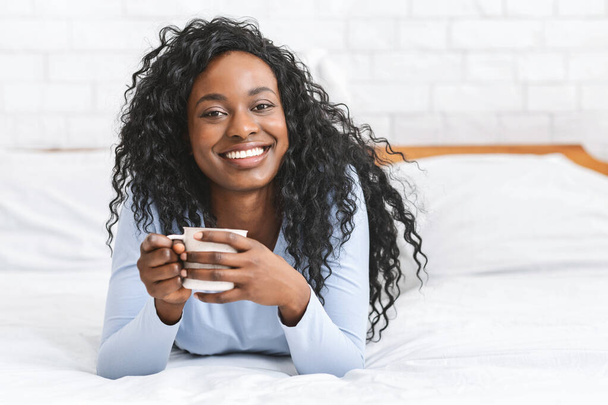 Χαρούμενο κορίτσι που χαλαρώνει στο κρεβάτι με ένα φλιτζάνι καφέ - Φωτογραφία, εικόνα