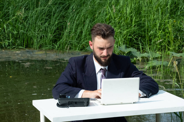 Office in the swamp. A young businessman with a beard in a suit as a clerk or manager sits in a quagmire at a white table and works on a laptop. Concept of freelance, crisis or routine. - Foto, Imagen
