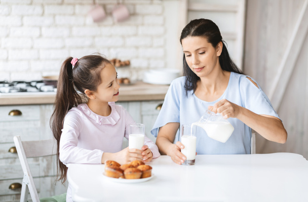 Mutter gießt Milch ins Glas, sitzt mit Tochter in Küche - Foto, Bild