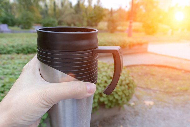 Season, autumn hot drinks and picnic concept - metal thermo coffee cup in woman hand sitting in park, coffee on the go - Foto, Bild
