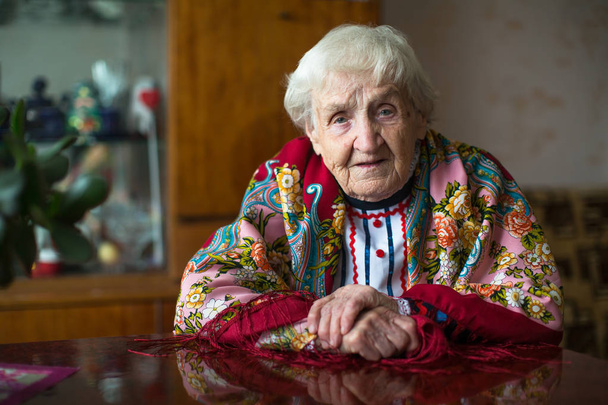 Γριά γυναίκα με φωτεινά ρούχα χειρονομίες κάθεται στο τραπέζι. - Φωτογραφία, εικόνα