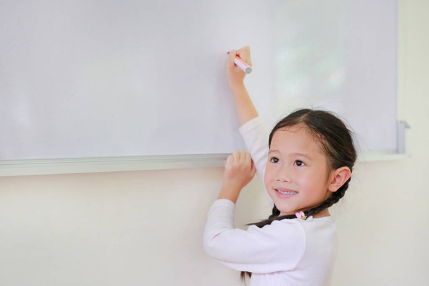 Portrait of Smiling little Asian child girl or Schoolgirl writing something on whiteboard with a marker and looking up in the classroom. White board with copy space for text. - Valokuva, kuva