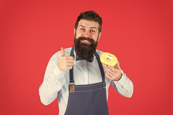 Cafe and bakery concept. Sweet donut from baker. Man bearded baker in cooking apron hold cute dessert. Ways to reduce hunger and appetite. Hipster bearded baker hold glazed donut on red background - Fotó, kép