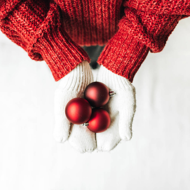 Boże Narodzenie / Nowy rok / Boże Narodzenie kompozycja. Młoda kobieta w czerwonym swetrze z dzianiny i białe rękawiczki trzymające zabawki choinkowe. Płaskie łóżko, widok z góry. Koncepcja minimalnego urlopu zimowego. - Zdjęcie, obraz