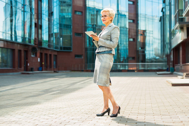 stylish adult woman in age in a suit and glasses against the background of a glass building with a tablet in her hands - Foto, Imagem