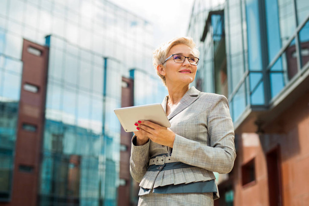 stylish adult woman in age in a suit and glasses against the background of a glass building with a tablet in her hands - Foto, imagen