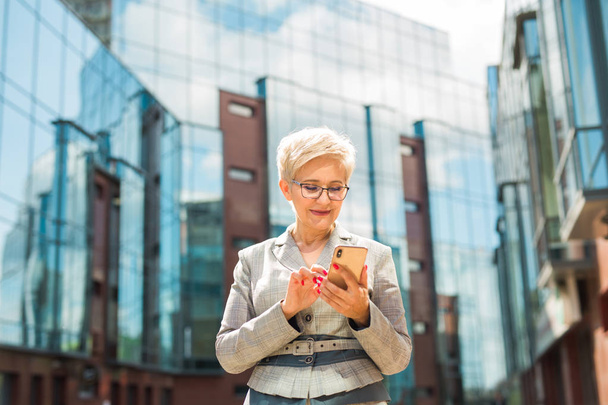 mulher adulta elegante na idade em um terno e óculos contra o fundo de um edifício de vidro
 - Foto, Imagem