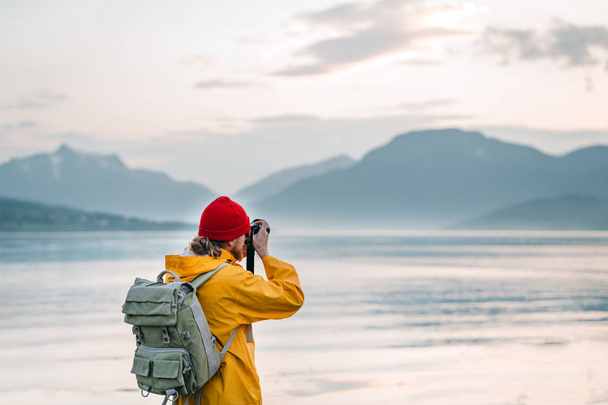 Traveler photographer wearing yellow raincoat taking photo by professional camera of fantastic scandinavia nature. Man tourist tourist travels to beautiful mountain and fjord places - Фото, зображення