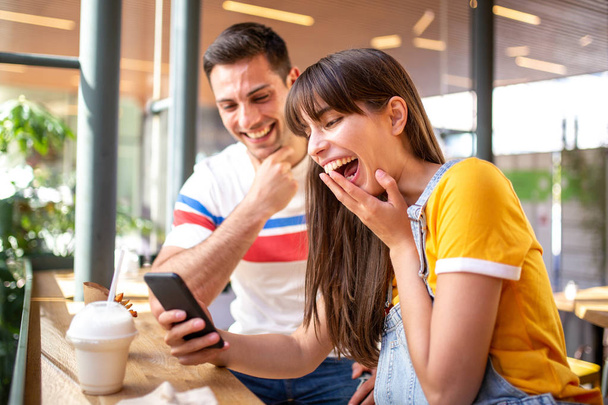Portrait of young couple laughing and looking at mobile phone at cafe - 写真・画像