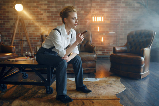 Γυναίκα με αυστηρά ρούχα κάθεται στο τραπέζι και το κάπνισμα πούρο, ρετρό μόδα, στυλ γκάνγκστερ. Vintage επιχειρηματίας κυρία στο γραφείο με τοίχους από τούβλα - Φωτογραφία, εικόνα