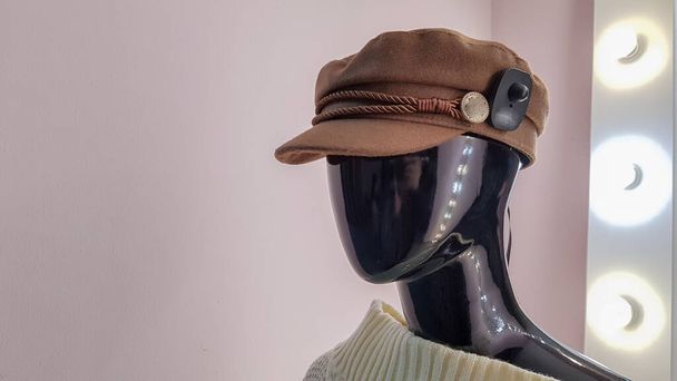 Portrait of a black mannequin with a brown hat in a women fashion store. Mannequin woman in a hat on a pink wall background and a large white mirror with backlight and light bulbs - 写真・画像