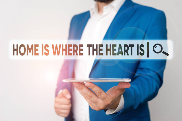 Text sign showing Home Is Where The Heart Is. Conceptual photo Your house is where you feel comfortable and happy Man in the blue suite and white shirt holds mobile phone in the hand. - Photo, Image