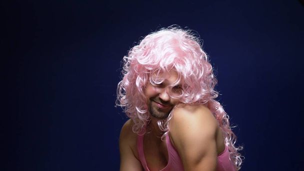 crazy handsome young guy in a curly wig and a pink t-shirt against a dark background is dancing funny, shows his muscles - Foto, Imagem