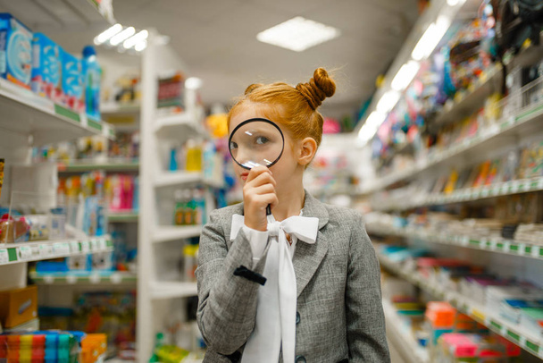 Μικρό κορίτσι του σχολείου κοιτάζει μέσα από μεγεθυντικό φακό, ψώνια σε χαρτοπωλείο - Φωτογραφία, εικόνα