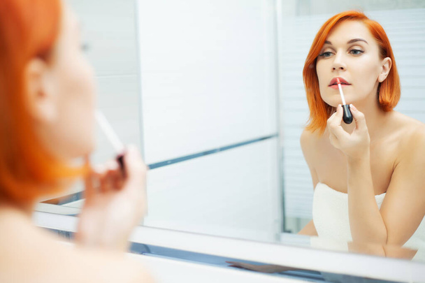 Bőrápolás. Egy vörös hajú nő sminkeli magát a világos fürdőszobában.. - Fotó, kép