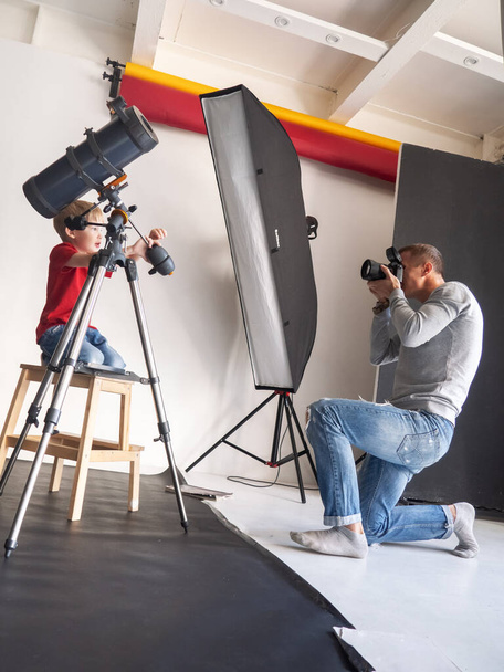 A man takes a portrait of a boy with a telescope in the studio with artificial light on a black background. - Fotoğraf, Görsel