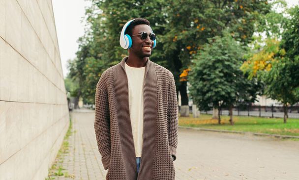 Cool modern african man in wireless headphones listening to music wearing brown knitted cardigan walking in autumn city park in rain - Фото, изображение