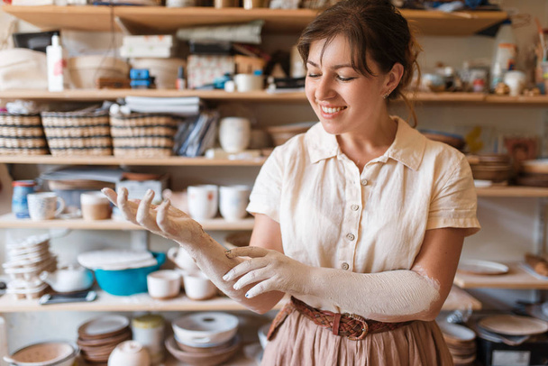 Female master hands covered with dried clay, pottery workshop interior on background. Woman molding a bowl. Handmade ceramic art, tableware making - Photo, Image