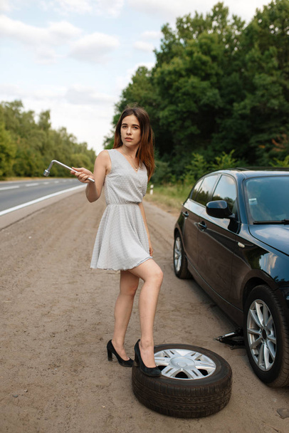Car breakdown, young woman puts the spare tyre. Broken automobile or problem with vehicle, trouble with punctured auto tire on highway - Photo, Image
