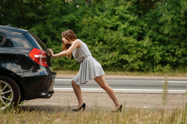 Kobieta pchająca zepsuty samochód na drodze, załamanie. Wypadek samochodowy lub awaryjny z pojazdem, problemy z silnikiem na autostradzie - Zdjęcie, obraz