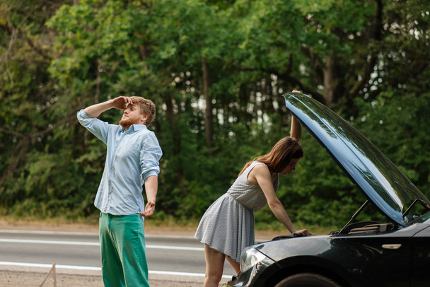 Tired couple at the opened hood on road, car breakdown. Broken automobile or emergency accident with vehicle, trouble with engine on highway - Photo, Image