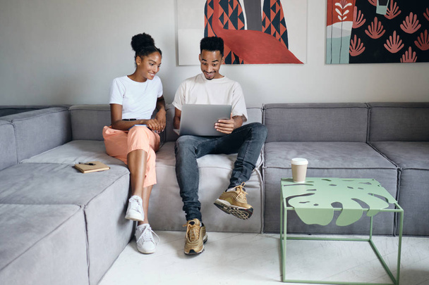 Genç ve çekici Afro-Amerikan çift modern evdeki kanepede keyifle birlikte dizüstü bilgisayarda çalışıyorlar. - Fotoğraf, Görsel