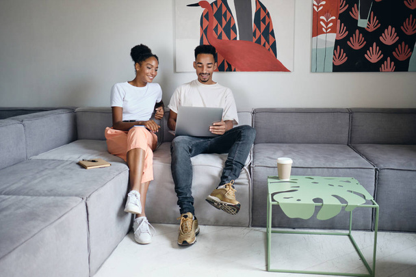 Νεαρό θετικό casual ζευγάρι Αφροαμερικανών ευτυχώς εργάζονται σε φορητό υπολογιστή μαζί σε καναπέ στο σύγχρονο σπίτι - Φωτογραφία, εικόνα