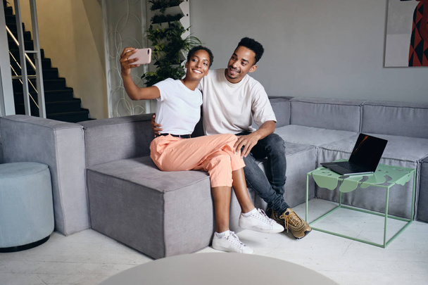 Νεαρό όμορφο casual African American ζευγάρι παίρνει ευτυχώς selfie στο κινητό μαζί στον καναπέ στο σύγχρονο σπίτι - Φωτογραφία, εικόνα