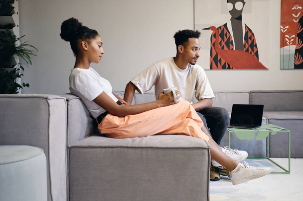 Jovem atraente casal afro-americano casual pensativo falando durante o coffee break no espaço de co-trabalho moderno
 - Foto, Imagem
