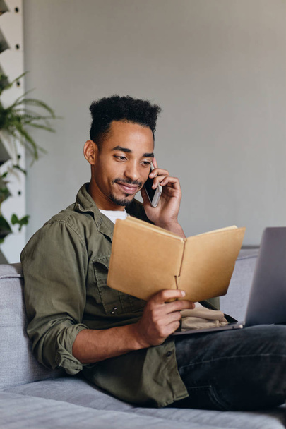 Fiatal vonzó mosolygós afro-amerikai férfi notebook boldogan beszél a mobiltelefon a modern co-working térben - Fotó, kép