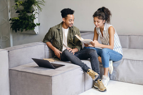Young African American man and pretty Asian woman happily reading book together in modern co-working space - Foto, immagini