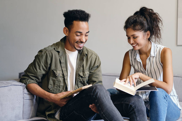 Joven hombre afroamericano sonriente y bonita mujer asiática leyendo alegremente un libro juntos en un moderno espacio de co-trabajo
 - Foto, Imagen