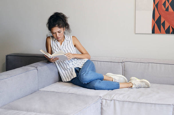 Красивая случайная азиатская девушка лежит на диване задумчиво читая книгу в современном доме
 - Фото, изображение