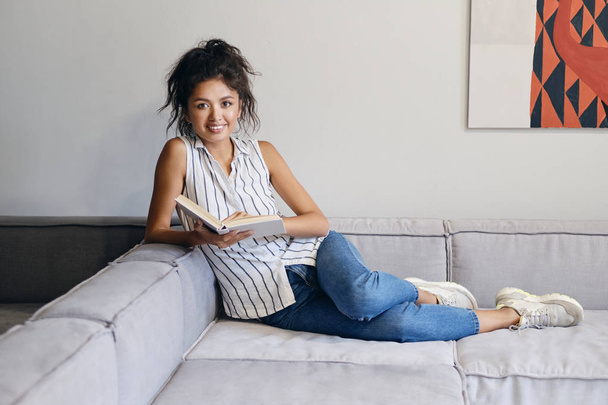 Bella allegra ragazza asiatica sdraiata sul divano con libro gioiosamente guardando in macchina fotografica a casa moderna
 - Foto, immagini