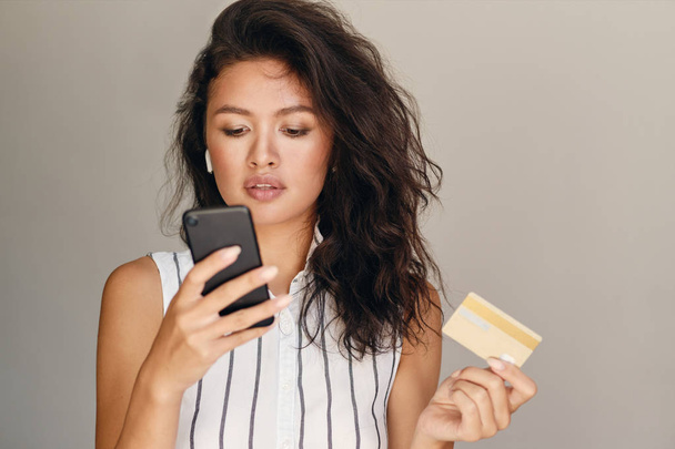 Портрет молодої вродливої азіатської жінки з кредитною карткою з упевненістю користується мобільним телефоном над сірим фоном. - Фото, зображення