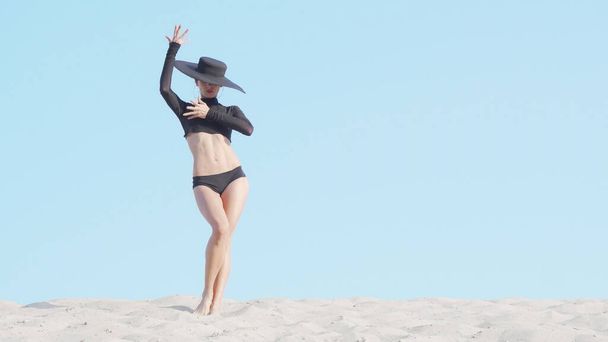 Εκπληκτική κομψή γυναίκα με μεγάλο μαύρο καπέλο χορεύει στην άμμο - Φωτογραφία, εικόνα