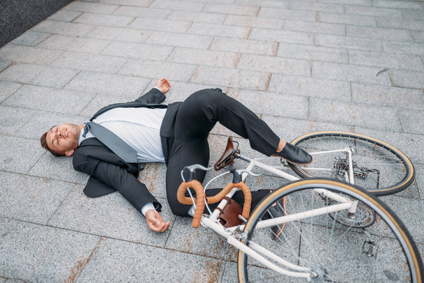 Businessman fell off his bike near office building in downtown. Business person riding on eco transport on city street - 写真・画像
