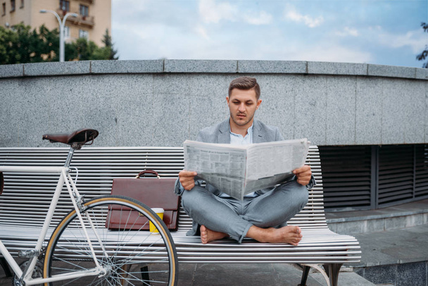 Επιχειρηματίας με ποδήλατο ανάγνωση εφημερίδα στον πάγκο στο κτίριο γραφείων στο κέντρο της πόλης. Επιχειρηματίας ιππασία στις οικολογικές μεταφορές στο δρόμο της πόλης, αστικό στυλ - Φωτογραφία, εικόνα